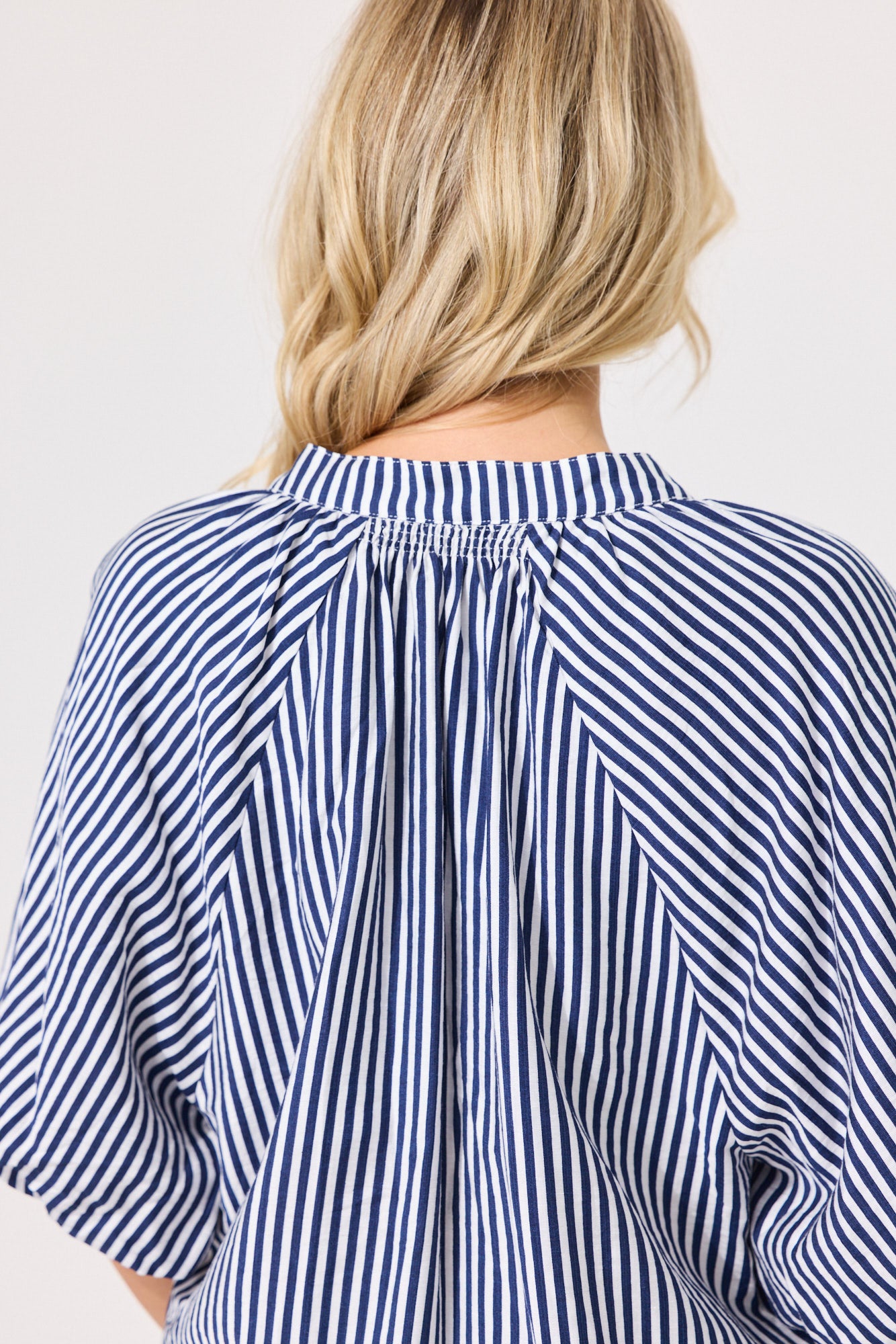 Back view of a woman wearing a navy and white vertically striped blouse with a gathered, pleated back and short wide sleeves, against a plain light gray background.