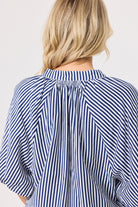 Back view of a woman wearing a navy and white vertically striped blouse with a gathered, pleated back and short wide sleeves, against a plain light gray background.