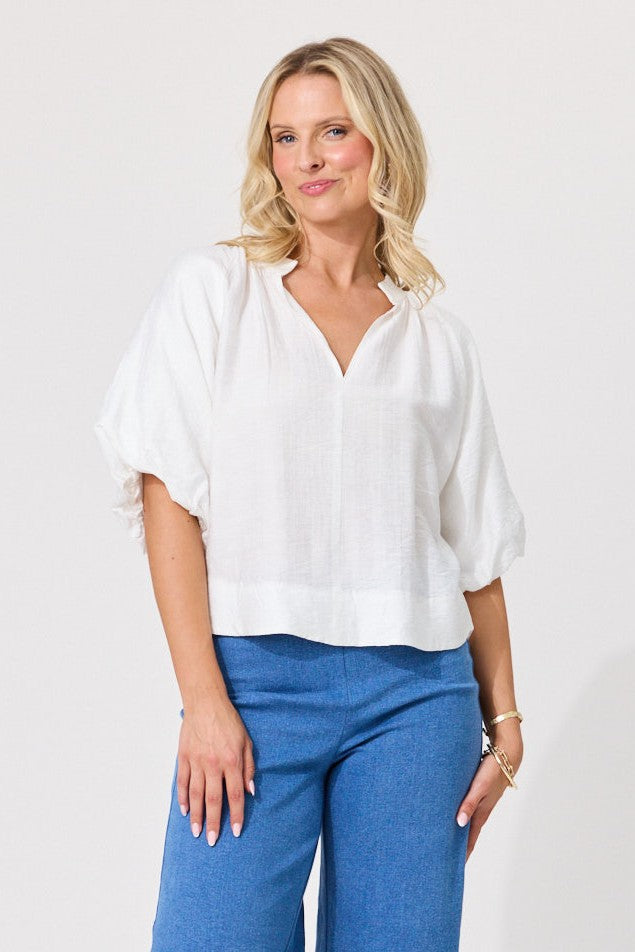 A woman wearing a white short-sleeve blouse with a V-neck and blue pants, standing against a plain light gray studio background.