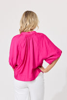 A woman viewed from the back wearing a bright hot pink blouse with voluminous elbow-length sleeves and white pants, set against a plain light gray studio background.