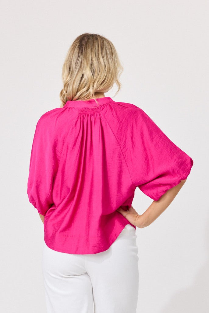 A woman viewed from the back wearing a bright hot pink blouse with voluminous elbow-length sleeves and white pants, set against a plain light gray studio background.