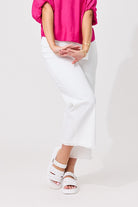 A model wearing white wide-leg cropped pants, a bright pink top, and white platform sandals with silver studs, standing against a plain white studio background.