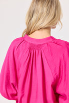 Close-up back view of a woman wearing a bright pink blouse with a gathered smocked neckline and voluminous sleeves, against a plain light background.