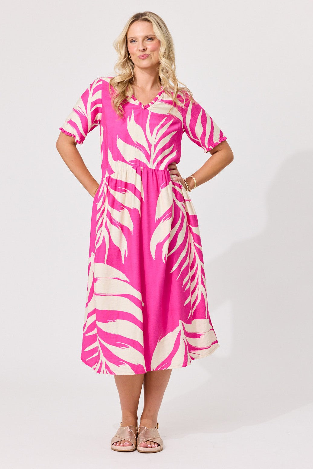 A woman modeling a short-sleeve midi dress with a vibrant pink and cream tropical leaf print, standing against a light gray studio background.