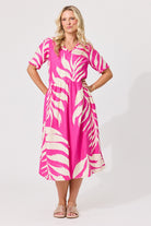 A woman modeling a short-sleeve midi dress with a vibrant pink and cream tropical leaf print, standing against a light gray studio background.