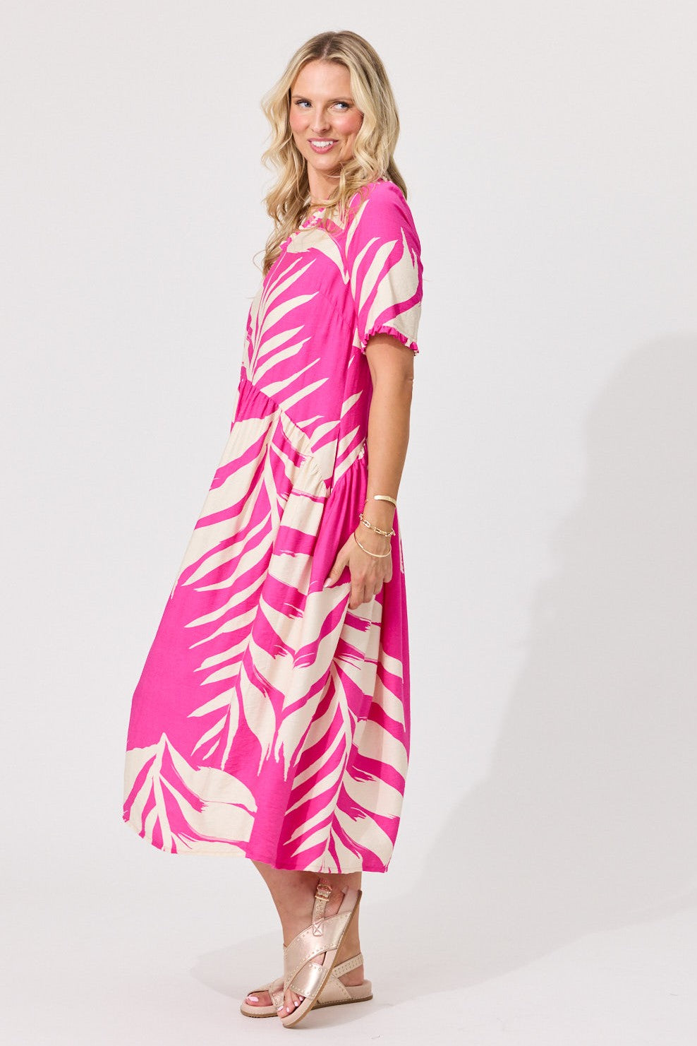 A woman modeling a short-sleeve midi dress with a bright pink and cream palm leaf pattern, standing against a plain light gray studio background.