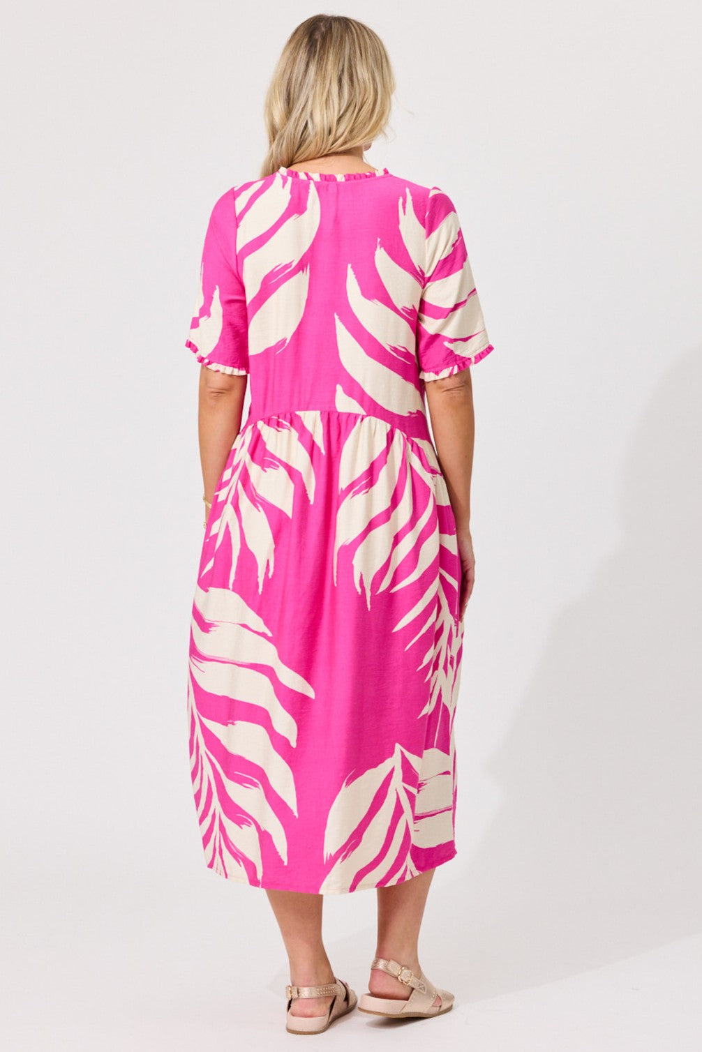 A woman viewed from the back wearing a bright pink midi dress with a large cream tropical leaf print, paired with beige sandals, against a plain white studio background.