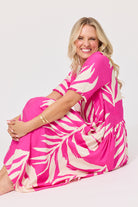 A smiling blonde woman sits cross-legged on a light gray studio floor, wearing a bright pink dress with a large off-white palm leaf print.