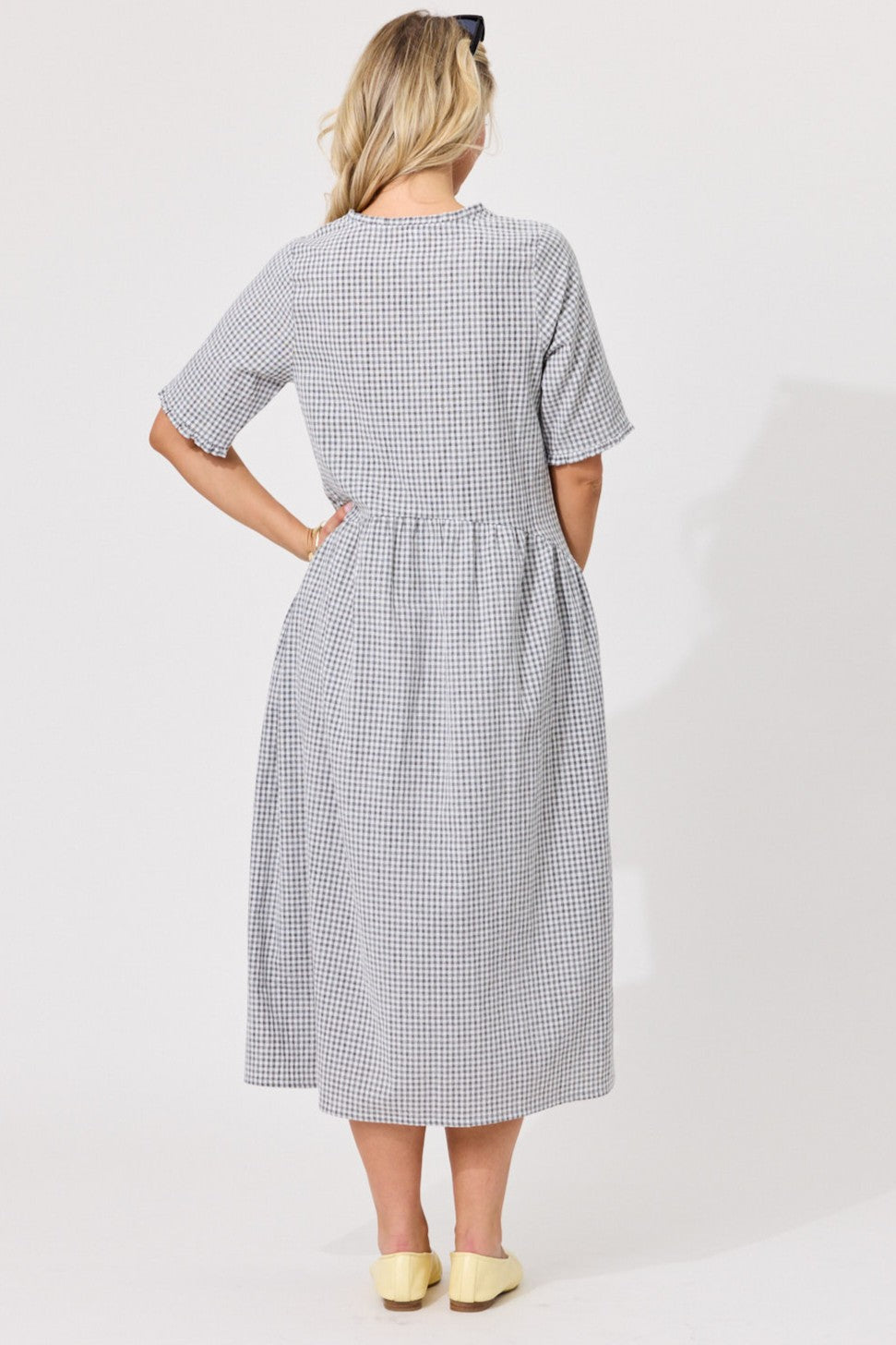 A woman viewed from the back wearing a short-sleeve midi dress with a small black and white gingham pattern, paired with pale yellow shoes against a plain light background.