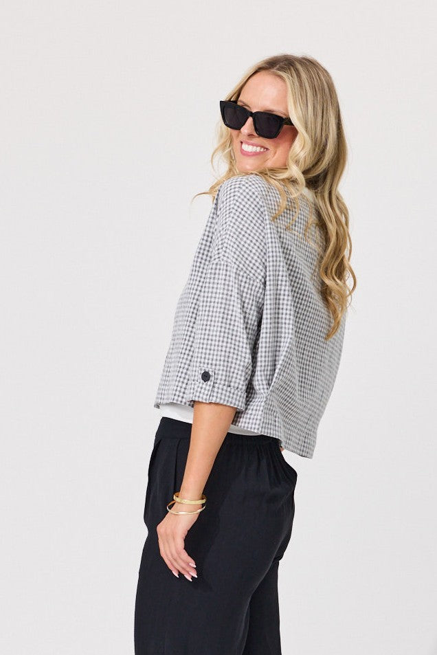 A woman wearing a black and white gingham cropped blouse with three-quarter sleeves and black pants, photographed against a plain light gray studio background.