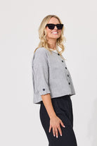 A woman modeling a cropped black and white gingham double-breasted jacket with three-quarter sleeves and black buttons, paired with black trousers and sunglasses, against a plain light gray background.