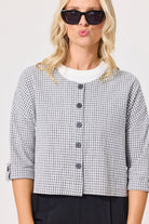 A woman wearing a cropped black and white gingham button-front jacket over a white crewneck top, styled with black sunglasses and dark trousers against a light gray studio background.
