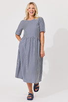 A woman modeling a navy and white vertically striped midi dress with short puff sleeves, a round neckline, and a gathered waist, paired with dark blue slide sandals against a light gray studio background.