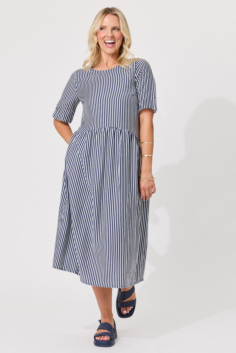 A woman modeling a navy and white vertically striped midi dress with short puff sleeves, a round neckline, and a gathered waist, paired with dark blue slide sandals against a light gray studio background.