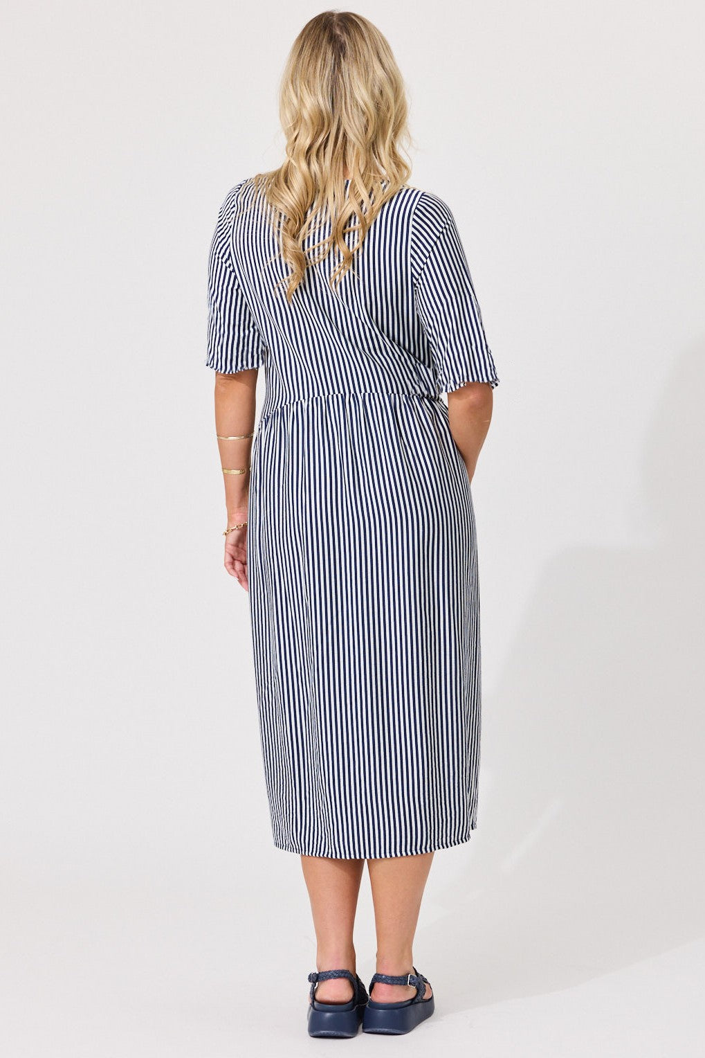 A woman viewed from the back wearing a navy and white vertically striped midi dress with short sleeves and side pockets, paired with dark blue Mary Jane shoes, against a plain white studio background.
