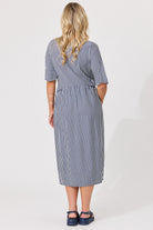 A woman viewed from the back wearing a navy and white vertically striped midi dress with short sleeves and side pockets, paired with dark blue Mary Jane shoes, against a plain white studio background.