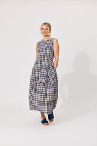 A woman modeling a sleeveless black and white gingham maxi dress with pockets, standing against a plain light gray studio background.