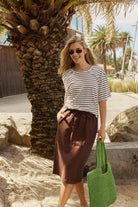 A woman wearing a black and white striped short-sleeve t-shirt and a brown midi skirt stands outdoors next to a palm tree, holding a green woven tote bag.