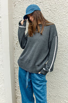 Person wearing a dark gray sweater with white accents and blue jeans against a light gray wall.
