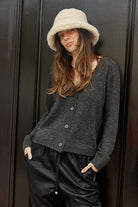 Woman wearing a gray cardigan, black pants, and a beige bucket hat against a dark background