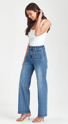 A woman modeling a pair of high-waisted, wide-leg blue jeans with a white tank top and beige heeled sandals, standing against a plain white studio background.