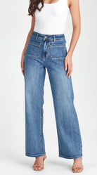 A model wearing a pair of high-waisted, wide-leg blue denim jeans with a white tank top and strappy heels, shown from the waist down against a plain white studio background.