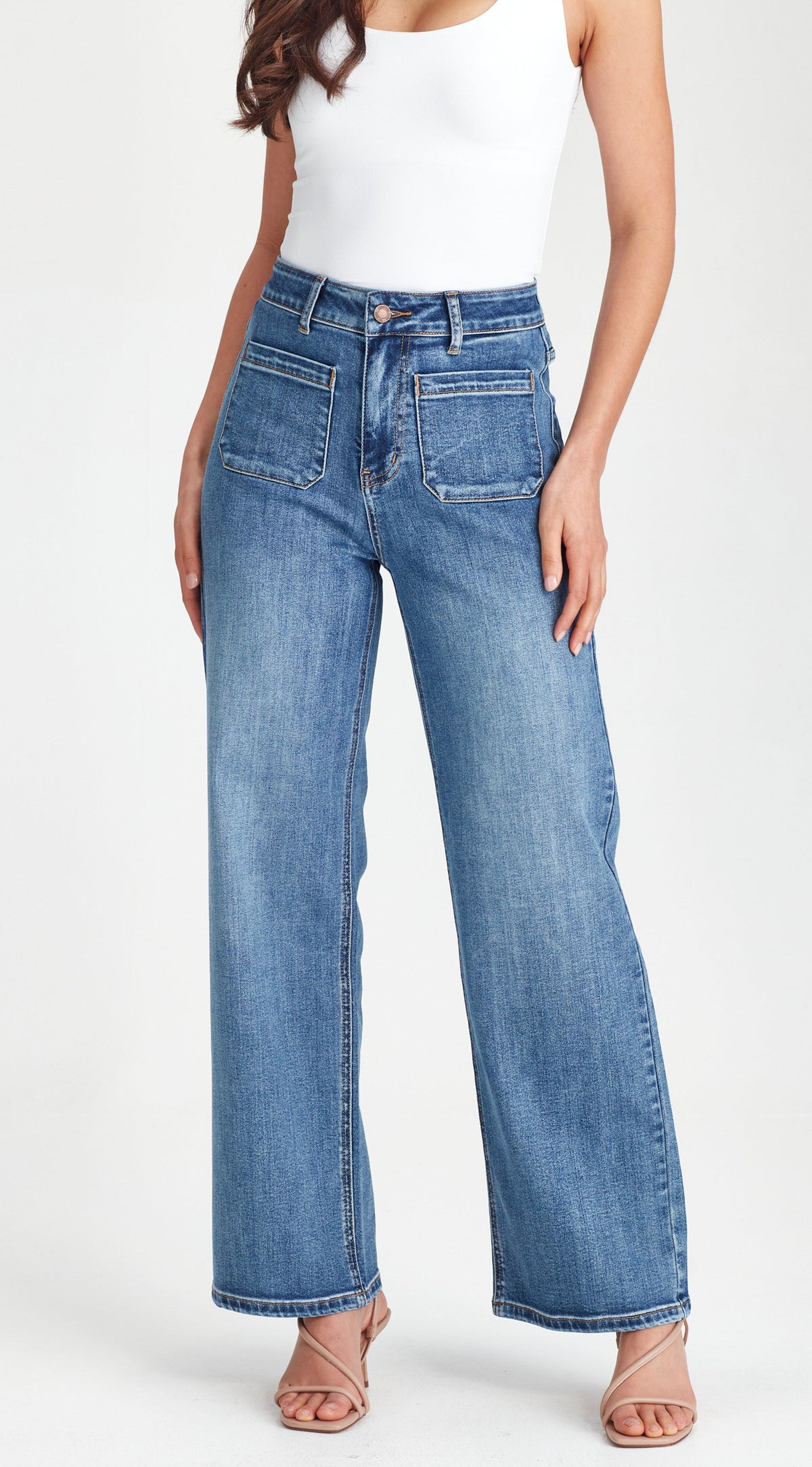 A model wearing a pair of high-waisted, wide-leg blue denim jeans with a white tank top and strappy heels, shown from the waist down against a plain white studio background.