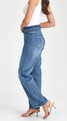 A woman wearing high-waisted, wide-leg blue denim jeans, a white sleeveless top, and beige high-heeled sandals, shown from a side-back angle against a plain white studio background.