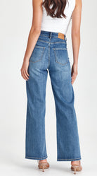 A woman shown from the back wearing high-waisted, wide-leg blue denim jeans with a white sleeveless top and beige heels, photographed against a clean white studio background.
