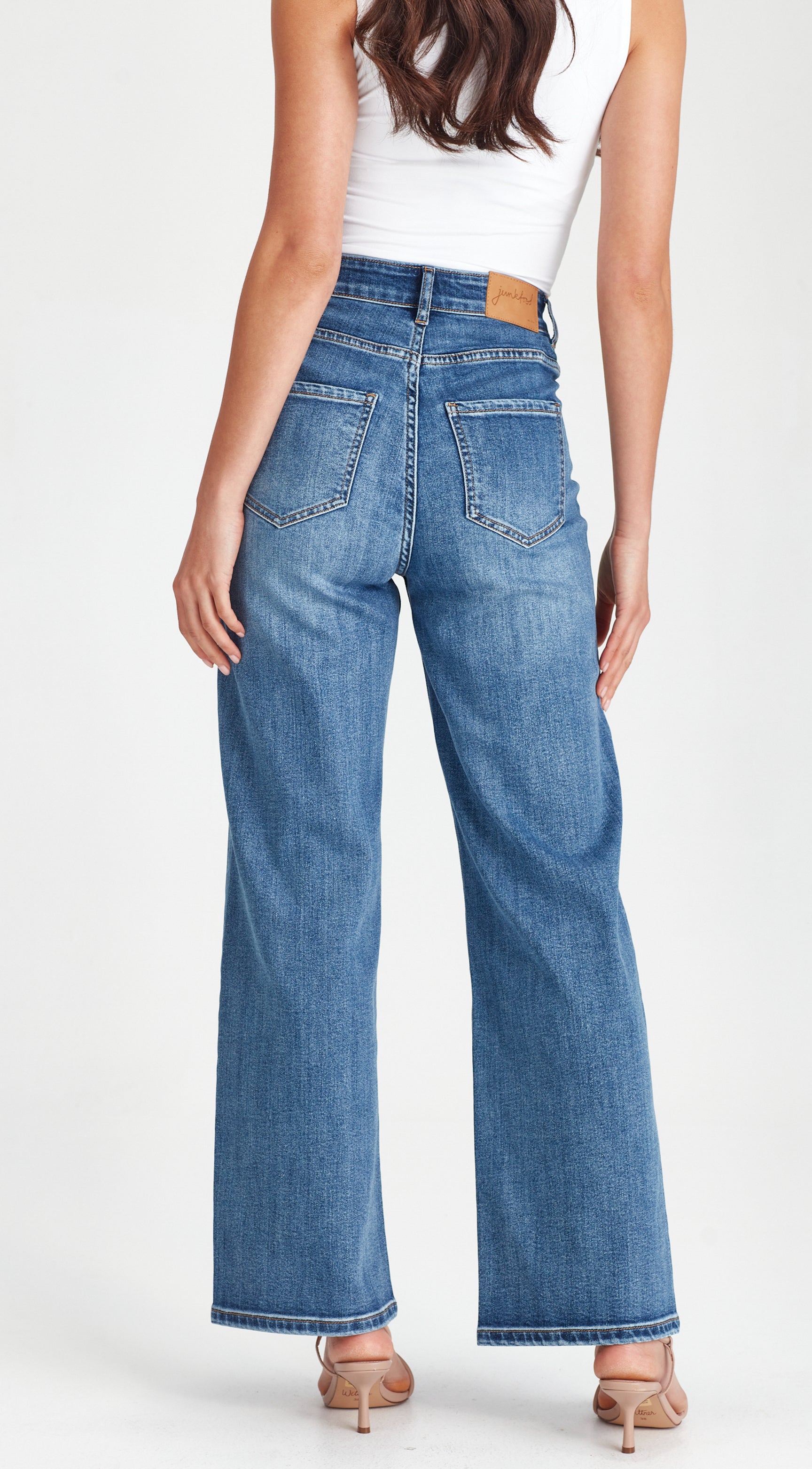 A woman shown from the back wearing high-waisted, wide-leg blue denim jeans with a white sleeveless top and beige heels, photographed against a clean white studio background.