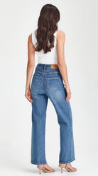 A woman viewed from the back wearing blue wide-leg jeans, a white sleeveless top, and nude high-heeled sandals against a light gray studio background.