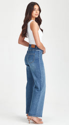 A woman modeling a pair of high-waisted, wide-leg blue jeans with a brown leather belt, paired with a white sleeveless top and nude high-heeled sandals, shown from a side-back angle against a plain white studio background.
