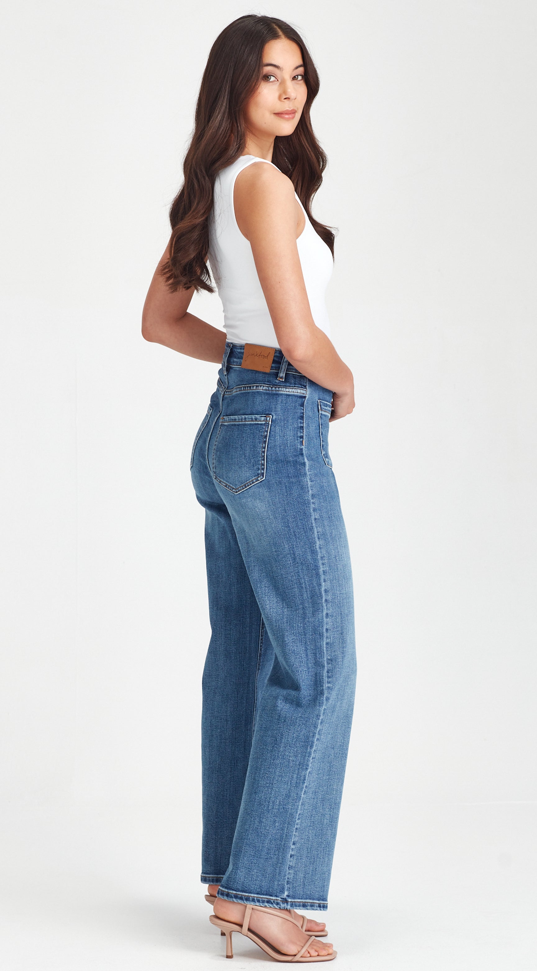 A woman modeling a pair of high-waisted, wide-leg blue jeans with a brown leather belt, paired with a white sleeveless top and nude high-heeled sandals, shown from a side-back angle against a plain white studio background.