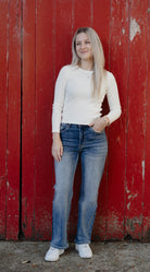 Person wearing a white long-sleeve shirt and blue jeans standing against a red wooden wall.