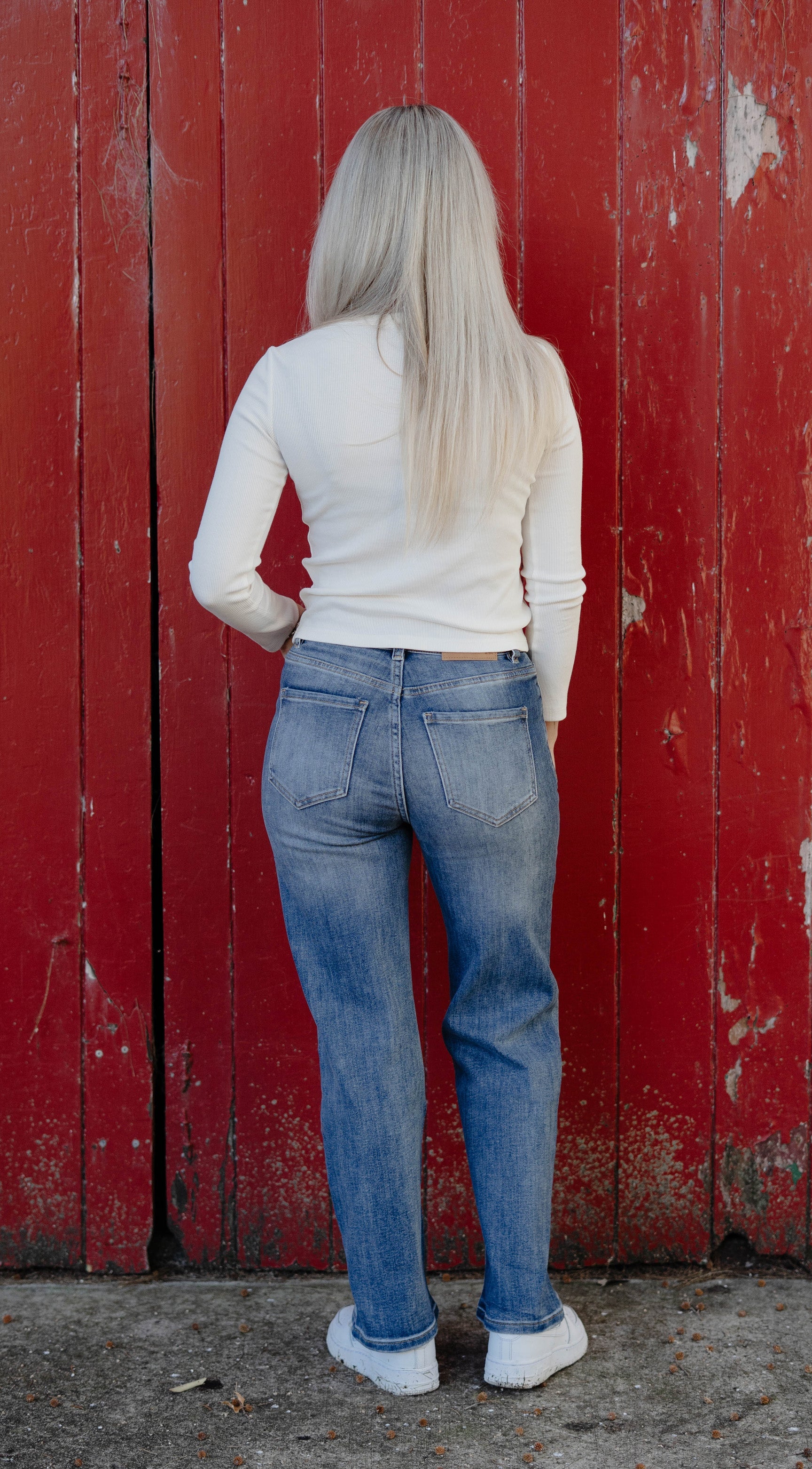 Person wearing blue jeans and a white long-sleeve shirt standing against a red wooden wall.