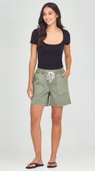 A woman modeling a black scoop-neck short-sleeve top and sage green drawstring cargo shorts with white sandals, standing against a light gray studio background.