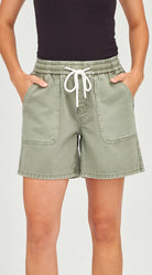 A person wearing light sage green denim shorts with a white drawstring waist, paired with a black top, shown from the chest to the upper thighs against a plain light gray background.