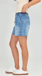 A person wearing light blue denim shorts with a drawstring waist, paired with a cream top and white sneakers, shown from the waist down against a light gray studio background.