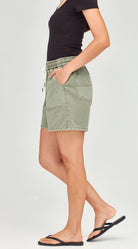 A model wearing light sage green denim shorts with a drawstring waist, paired with a black top and black flip-flops, shown from the waist down against a light gray studio background.