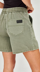 Close-up of a person wearing sage green denim shorts with an elastic waistband, back pocket, and a small black brand patch.