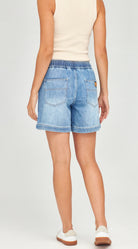 A model shown from the back wearing light-wash blue denim shorts with an elastic waistband, a cream sleeveless top, and white sneakers against a light gray studio background.
