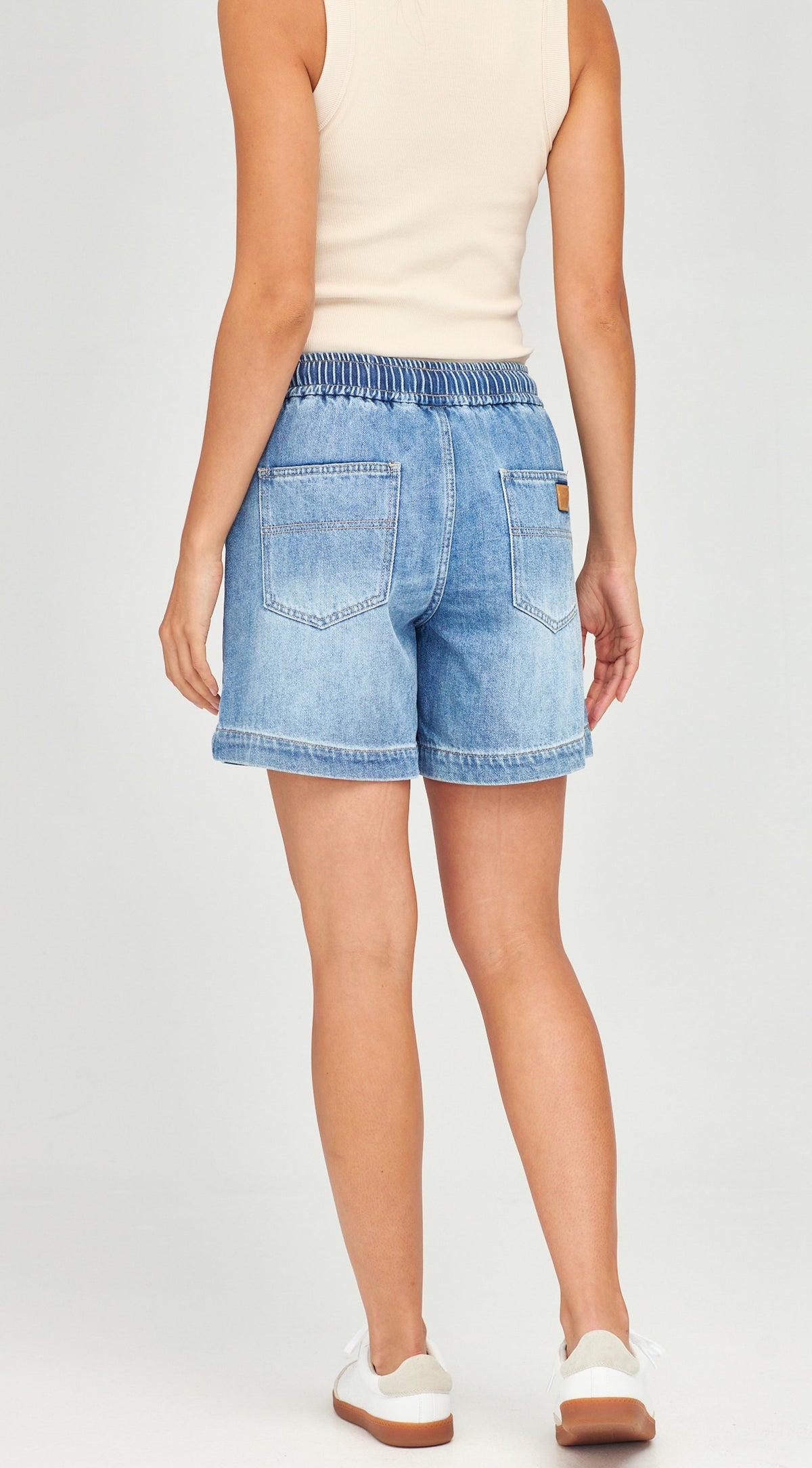 A model shown from the back wearing light-wash blue denim shorts with an elastic waistband, a cream sleeveless top, and white sneakers against a light gray studio background.