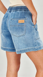 Close-up back view of a person wearing light blue denim shorts with an elastic waistband, back pockets, and a small tan brand patch on the upper right pocket.