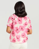 A woman viewed from behind wearing a short-sleeve blouse with a pink floral print on a cream background, paired with white pants, against a light gray studio background.