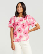 A woman modeling a short-sleeve blouse with a pink and white floral print, featuring ruffle details on the shoulders, paired with white pants against a light gray studio background.