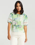 A woman modeling a short-sleeve patchwork blouse with green, white, and light blue floral prints, paired with off-white pants, against a light gray studio background.