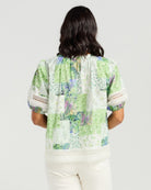 A woman viewed from behind wearing a short-sleeve blouse with a green, white, and purple botanical patchwork print, paired with cream pants, against a light gray studio background.