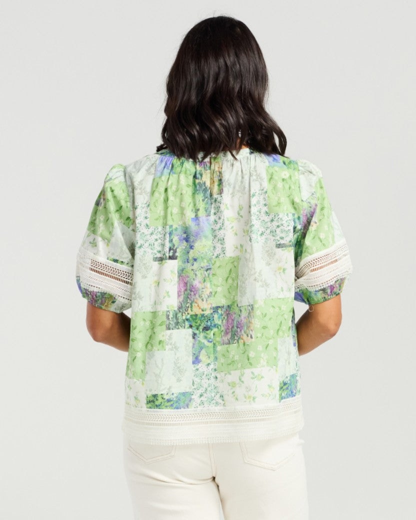 A woman viewed from behind wearing a short-sleeve blouse with a green, white, and purple botanical patchwork print, paired with cream pants, against a light gray studio background.