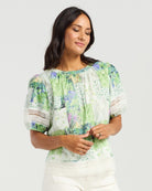 A woman wearing a short-sleeve blouse with a green, blue, and white abstract print, paired with white pants, photographed against a light gray studio background.