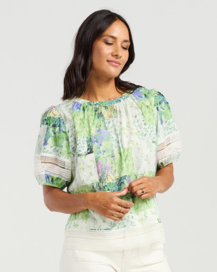 A woman wearing a short-sleeve blouse with a green, blue, and white abstract print, paired with white pants, photographed against a light gray studio background.